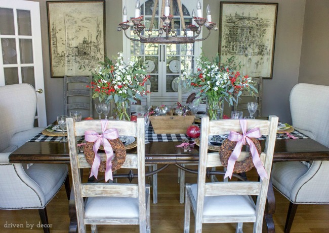 Dining room decorated for the holidays with a centerpiece of flowers and ornaments, star anise wreaths tied to the chair backs, jingle bells on chandelier, and pomegranate place cards