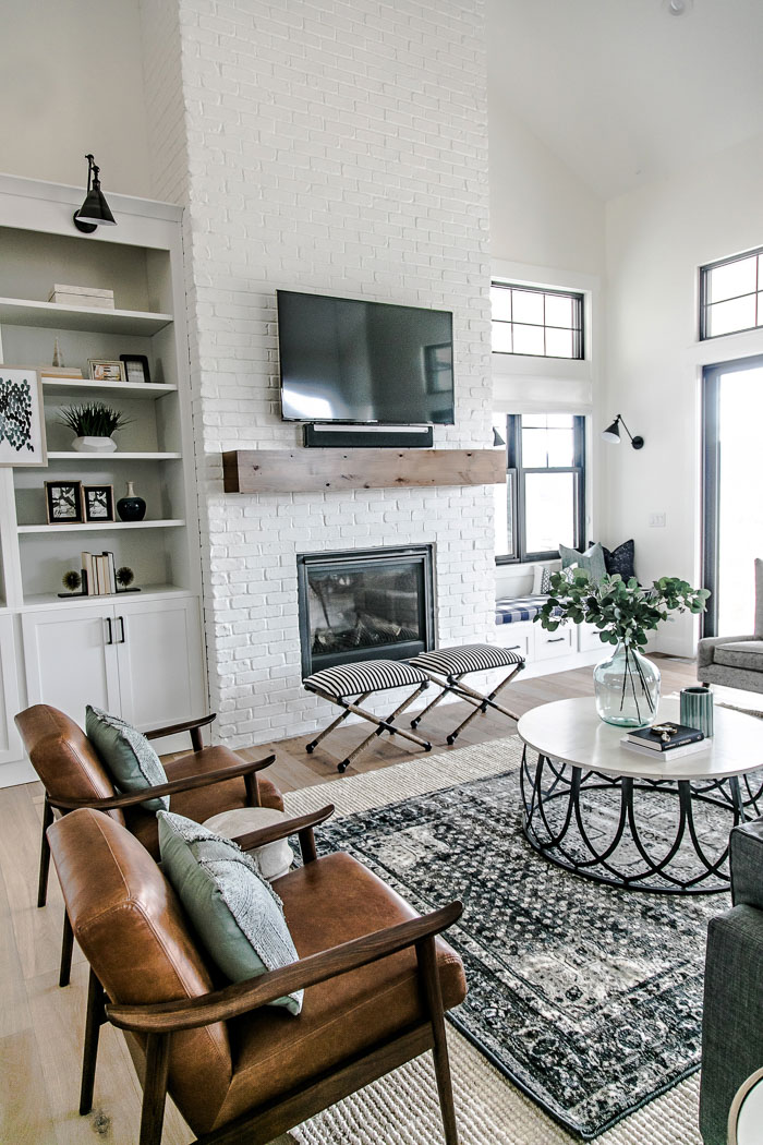 Gorgeous modern farmhouse living room designed by Sita Montgomery Interiors - white brick fireplace, simple wood mantel, leather slingback chairs, layered rugs, circular coffee table, and sconce lighting above open shelving