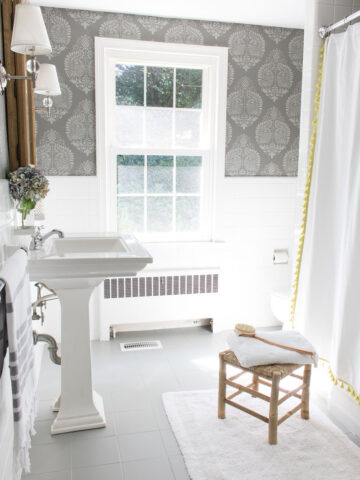 A budget bathroom remodel with ceramic tile floors painted gray and walls stenciled to look like wallpaper. Bathroom includes pedestal since, wood medicine cabinet, twin sconces, and tasseled shower curtain.