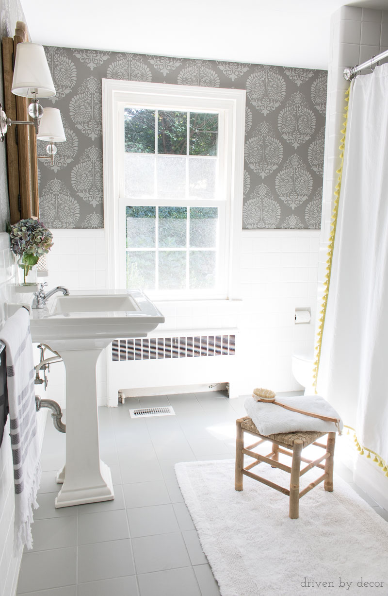 A budget bathroom remodel with ceramic tile floors painted gray and walls stenciled to look like wallpaper. Bathroom includes pedestal since, wood medicine cabinet, twin sconces, and tasseled shower curtain.