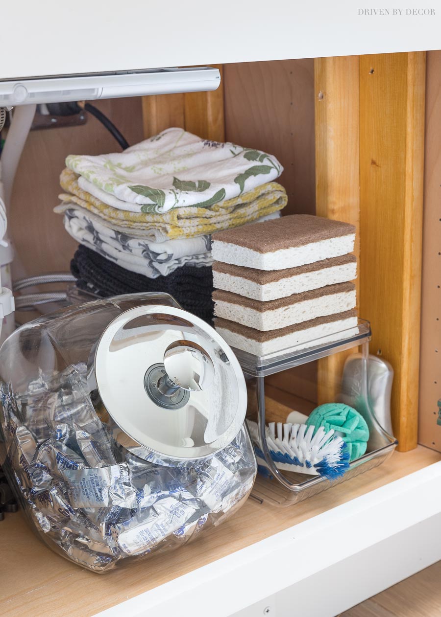 How I organize some of the space under our kitchen sink! A penny candy jar for detergent tabs and an acrylic organizer for scrub brushes, sponges, and towels!