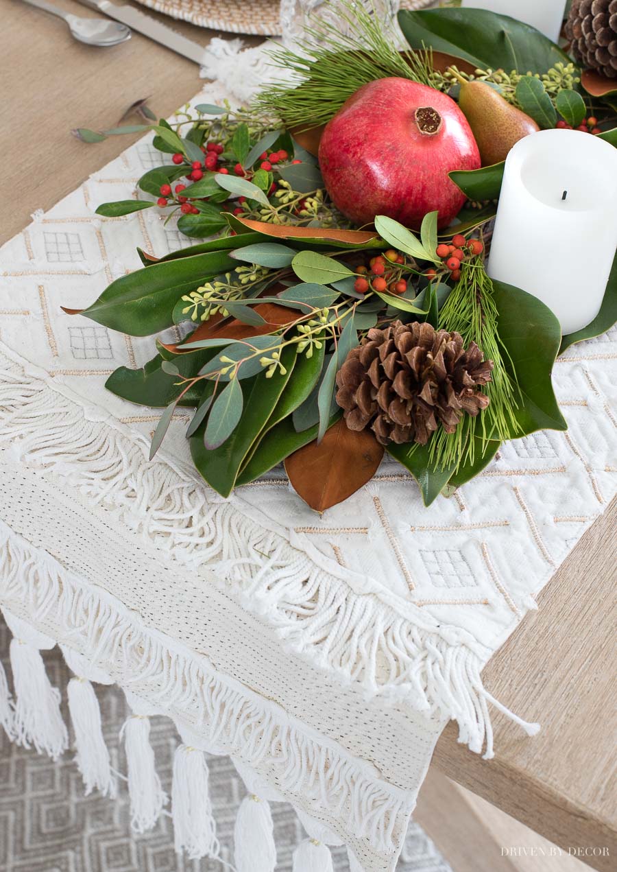 Gorgeous fringed table runner used as a base for a runner made of greenery, fruit, and pinecones.