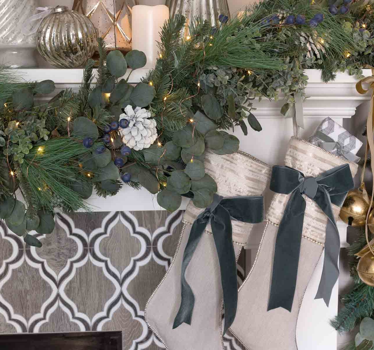 Our fireplace mantel decorated for Christmas with a greenery swag, stockings, jingle bells, and mercury glass trees
